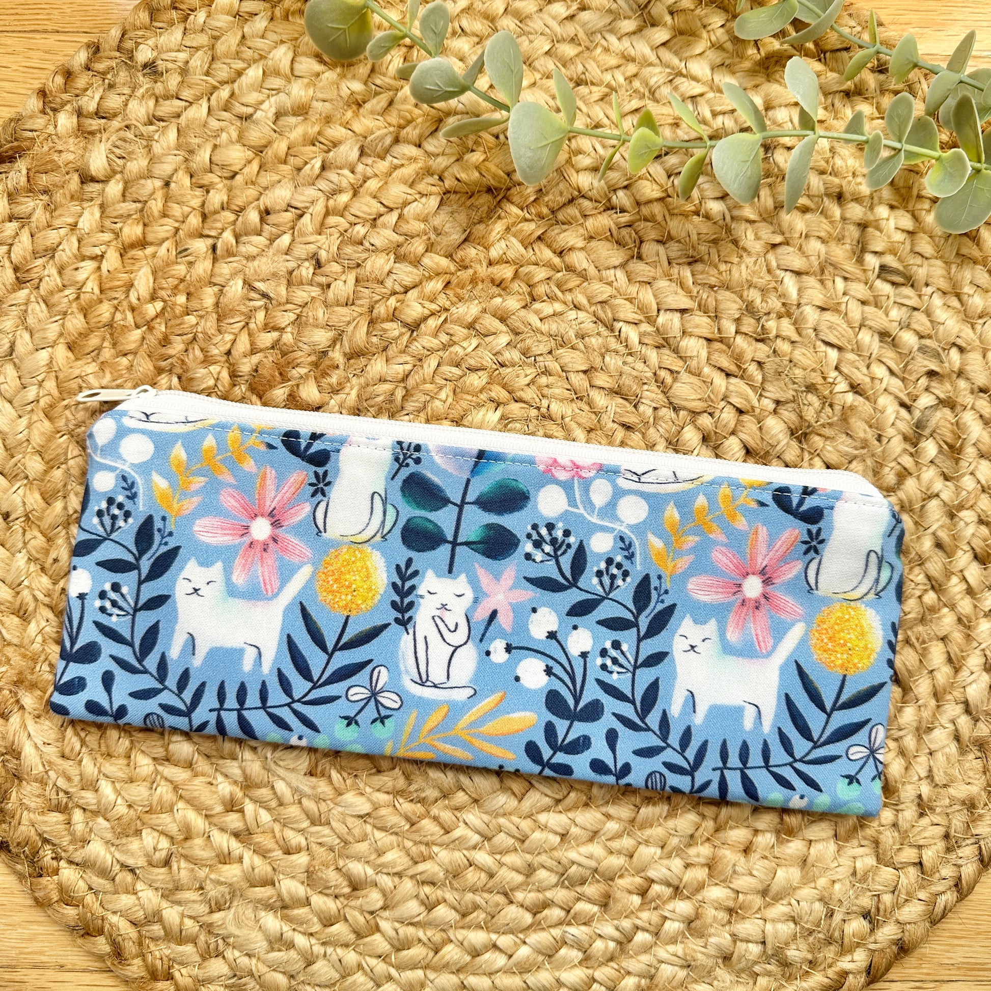 A rectangular zippered pouch with a blue background and a whimsical print of white cats, pink flowers, yellow pom-poms, and dark blue leaves.