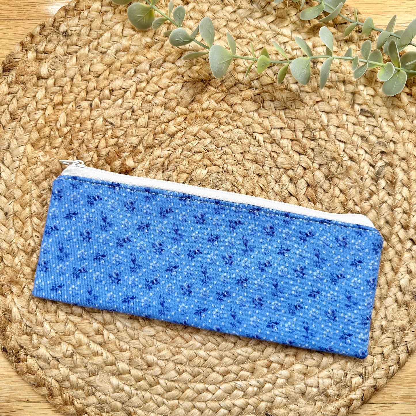 A blue fabric zippered pouch with a small white and dark blue floral pattern, resting on a round woven straw placemat with a sprig of green eucalyptus in the background.