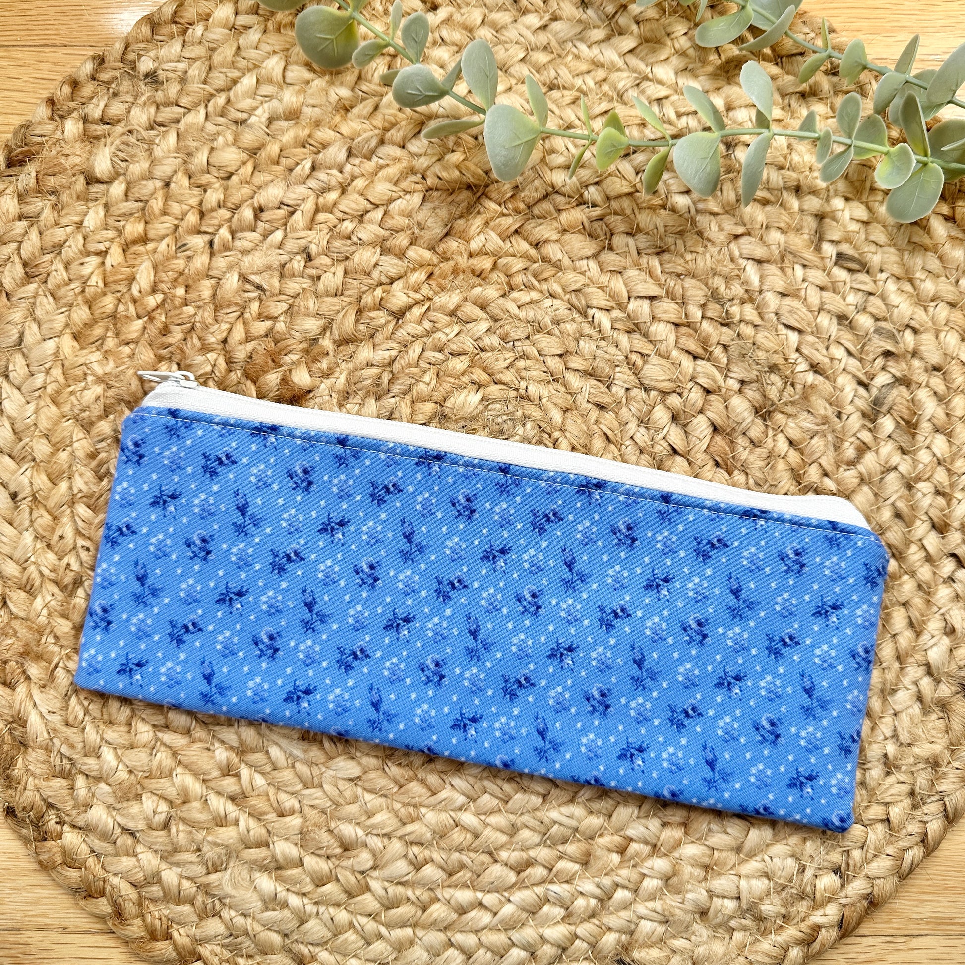 A blue fabric zippered pouch with a small white and dark blue floral pattern, resting on a round woven straw placemat with a sprig of green eucalyptus in the background.