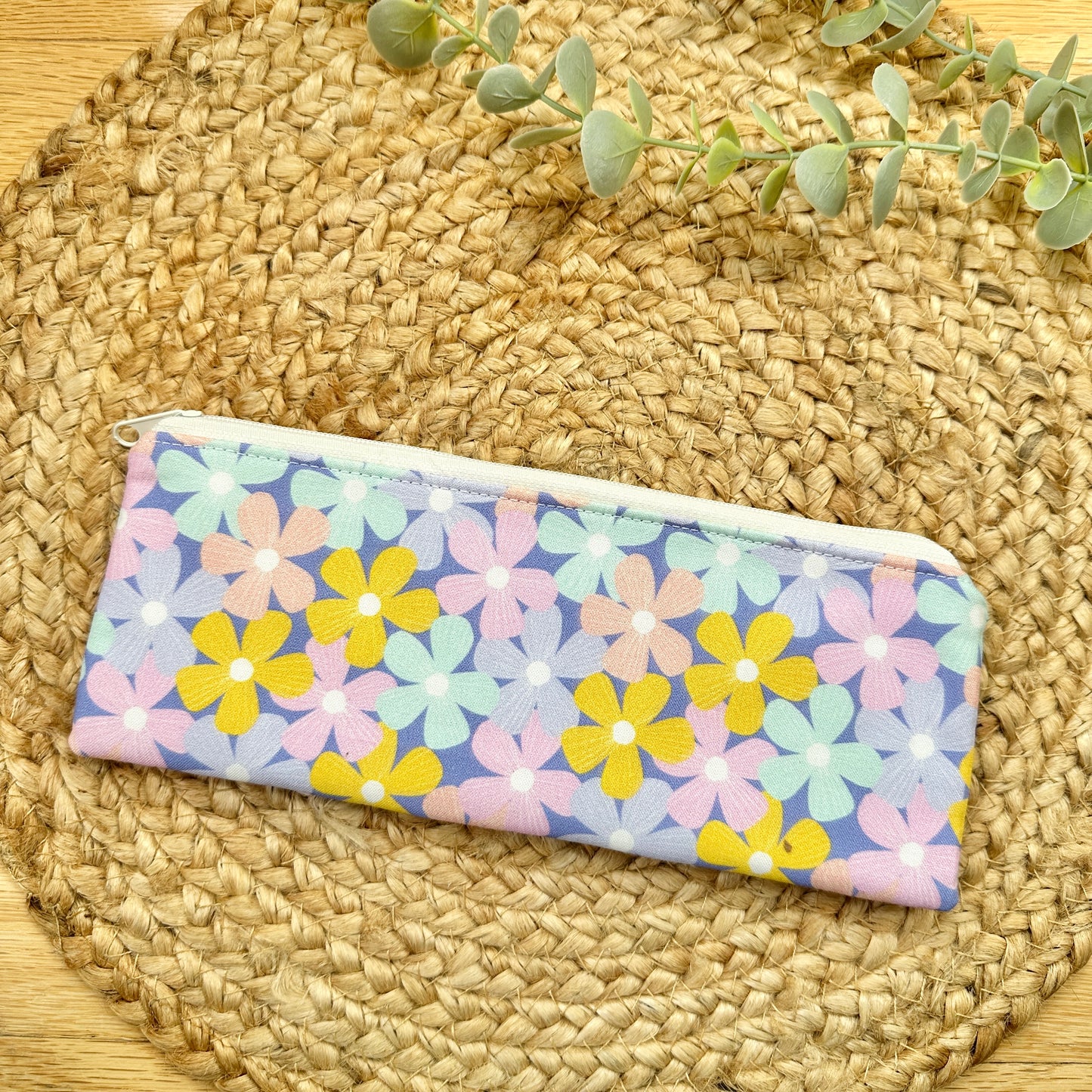A fabric zippered pencil case with a pastel floral print, displayed on a round woven straw placemat with a sprig of eucalyptus in the background.
