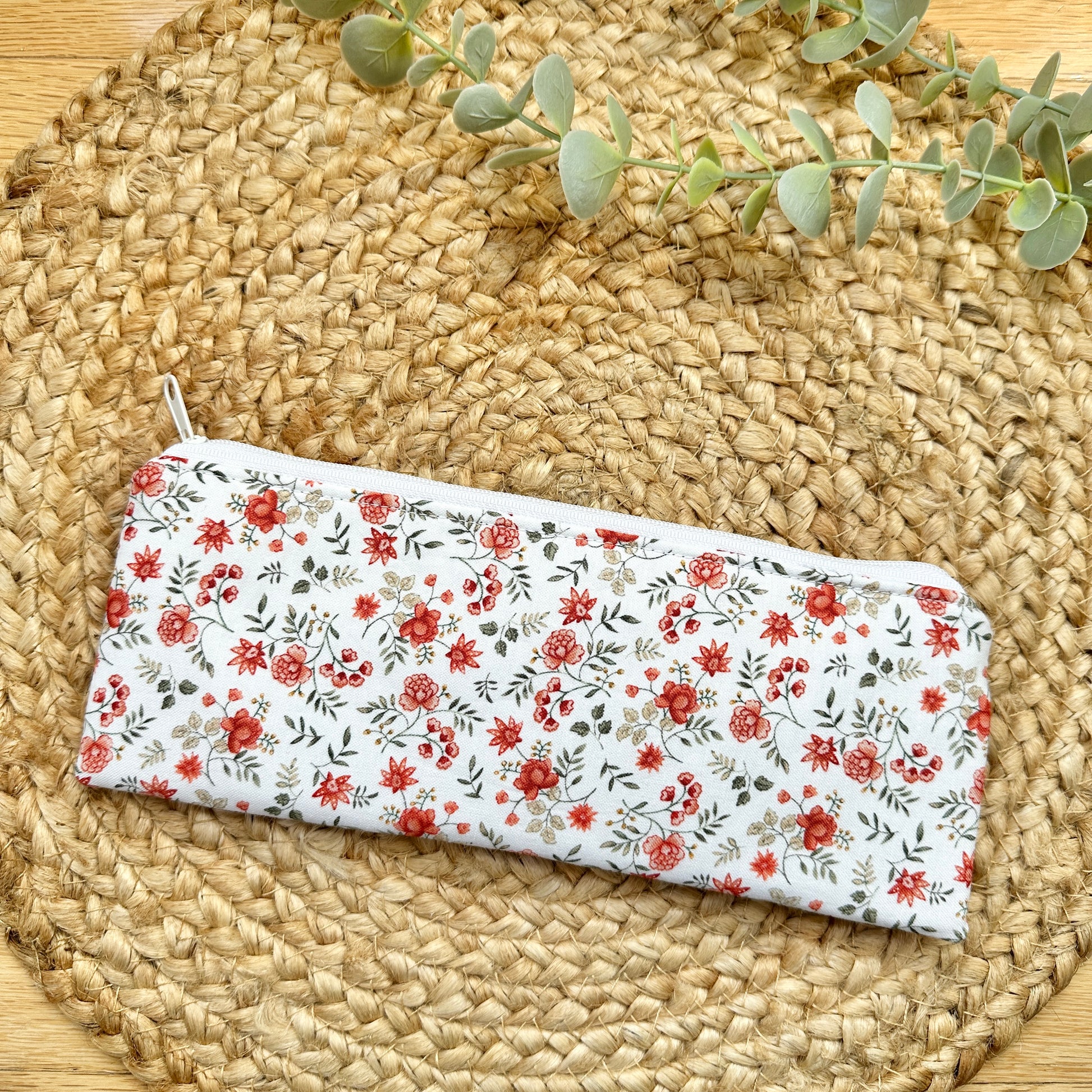 A rectangular zippered pouch with a white background and a red-orange and green floral print, displayed on a round woven straw mat with eucalyptus leaves.