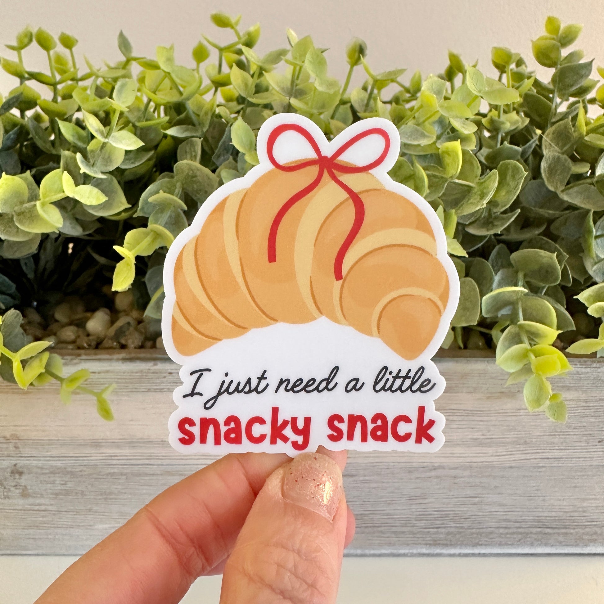 Sticker of a croissant with text 'I just need a little snacky snack' held in front of green foliage.