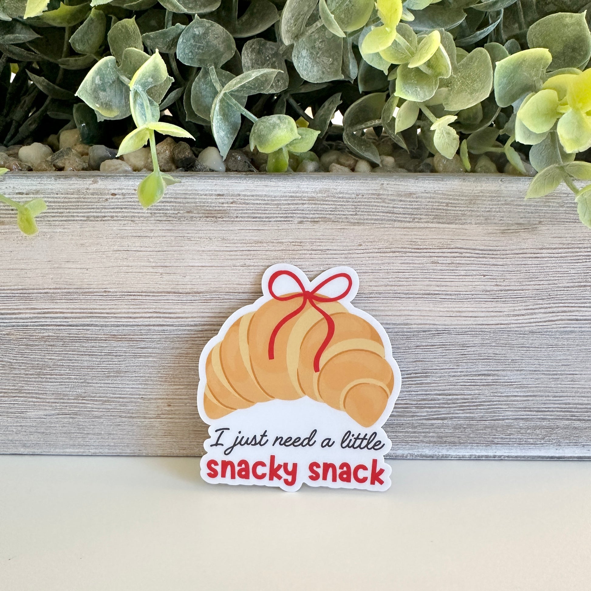 Sticker of a croissant with a bow and text 'I just need a little snacky snack' on a wooden surface with greenery.