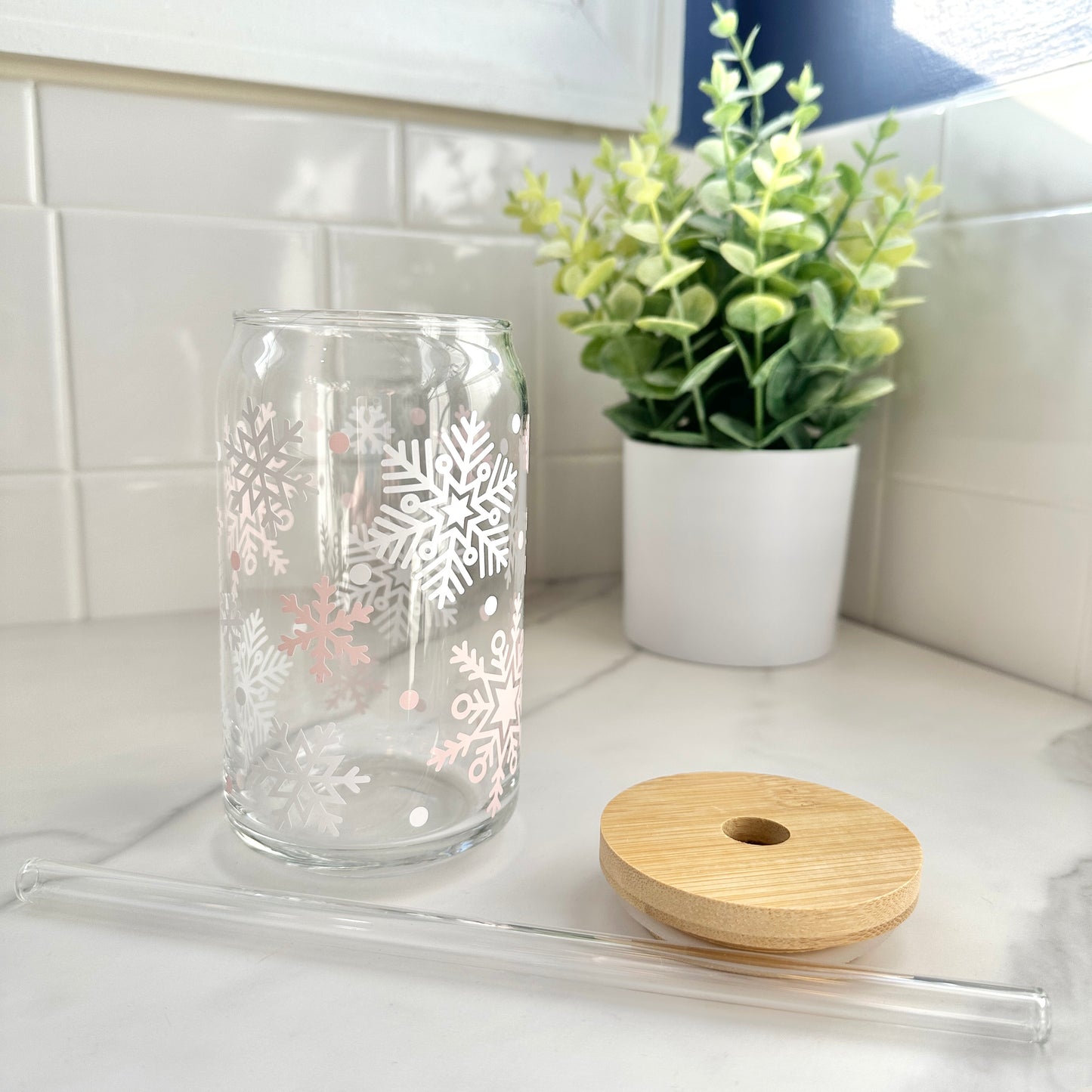Glass tumbler with snowflake design, wooden lid, and straw on a kitchen counter.