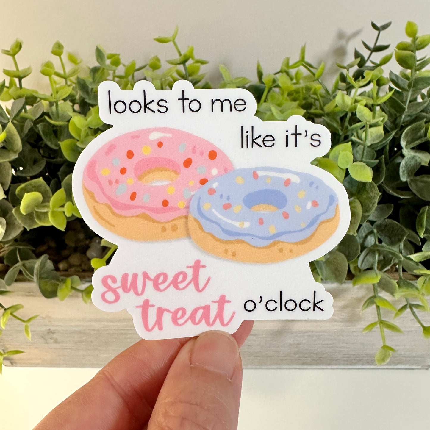 A hand holds a die-cut sticker featuring two illustrated donuts with pink and blue frosting and the text 'looks to me like it's sweet treat o'clock'. The background shows green artificial plants.