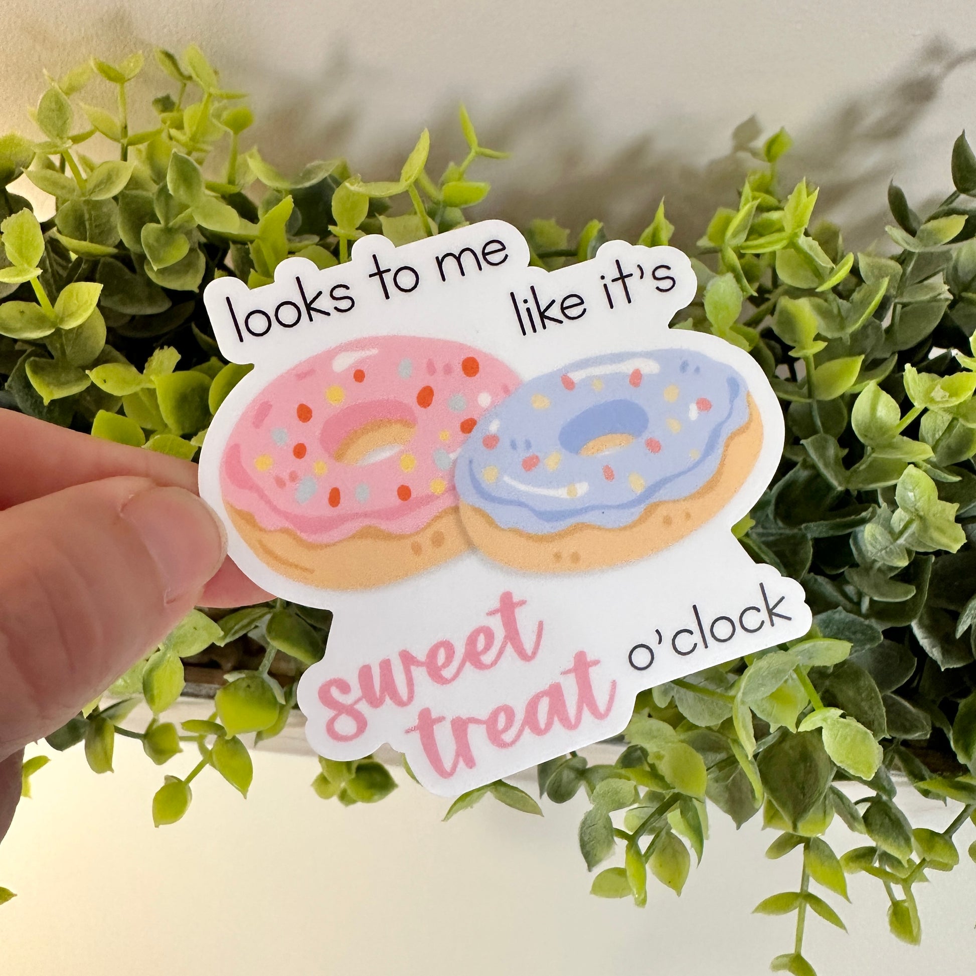 A hand holds a waterproof sweet treat sticker in front of a green plant.