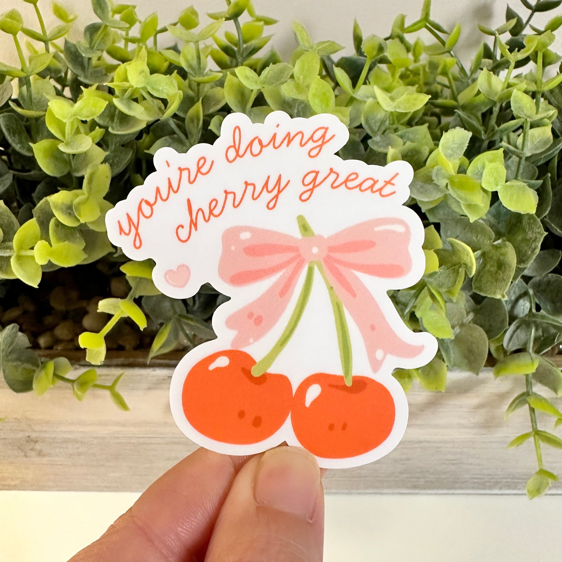 A hand holds a die-cut sticker featuring two cherries tied with a pink bow and the phrase 'you're doing cherry great' set against a background of green artificial foliage.