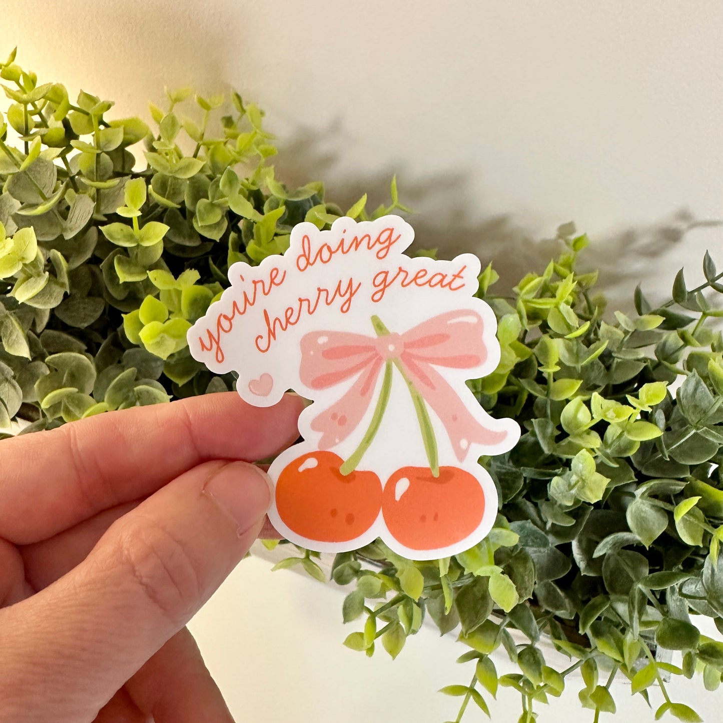 A hand holds a punny waterproof positivity decal in front of green artificial foliage.